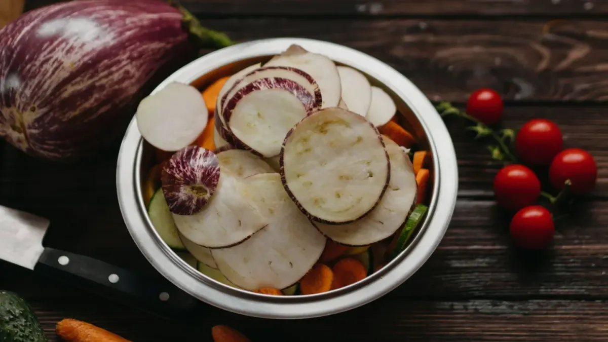 A dog's food bowl surrounded by fresh vegetables and a small pile of vitamin capsules